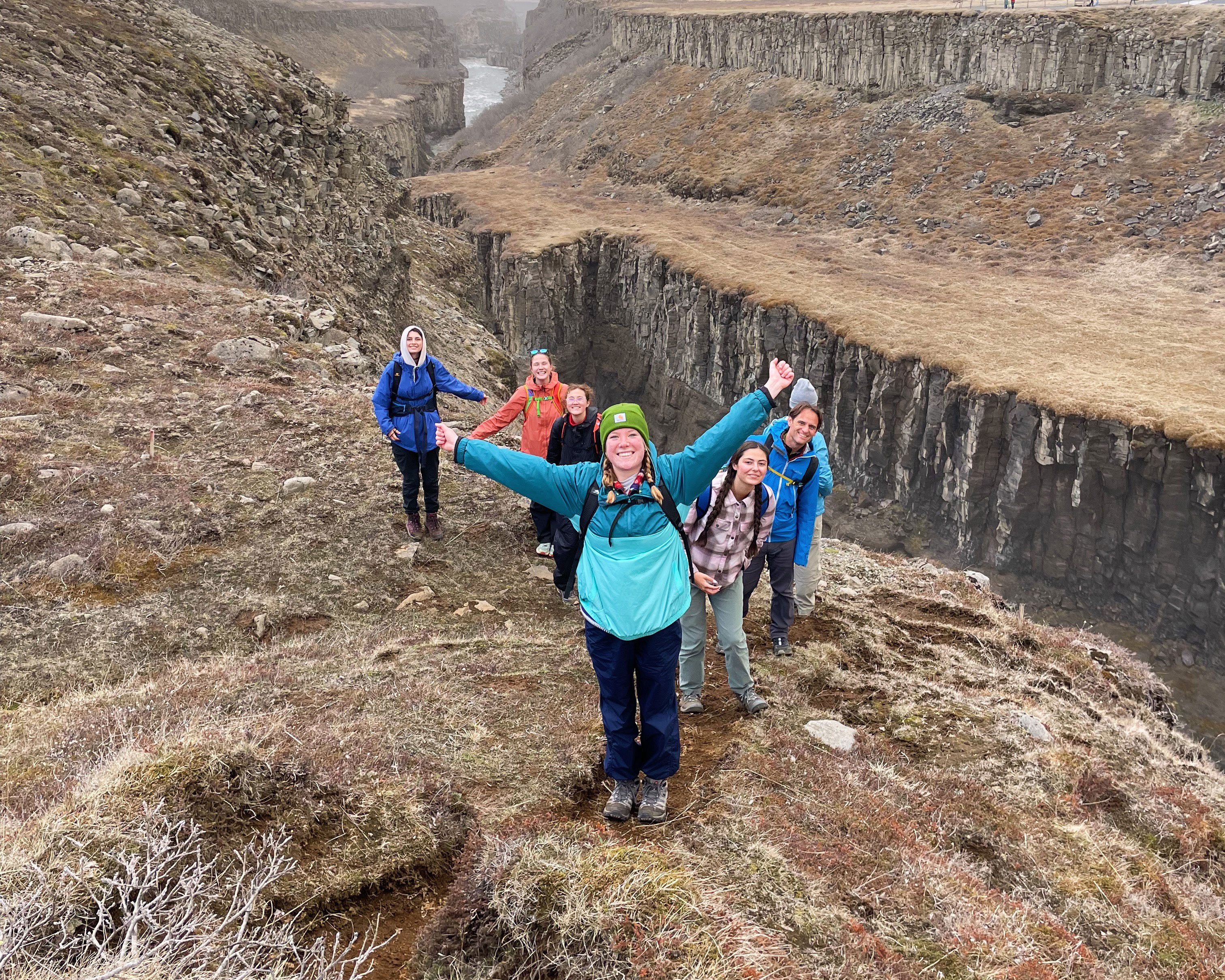 A group of hikers stands on a rocky trail in Iceland near the edge of a dramatic canyon with tall, rugged cliffs and a winding river far below.