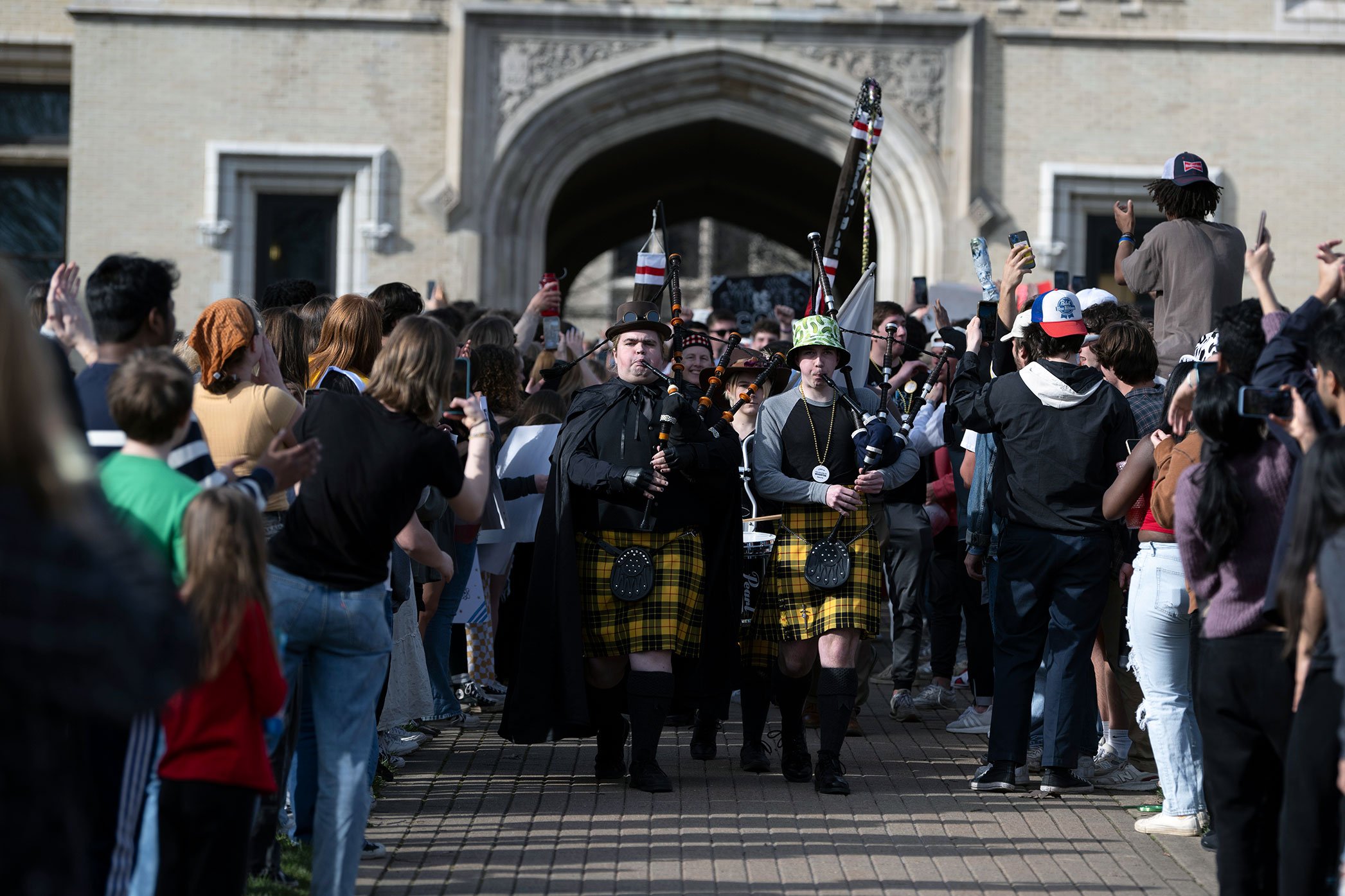 A crowd surrounds two bagpipers in yellow tartan kilts leading the I.S. Parade at The College of Wooster. They march outside a stone building with an arched entrance as onlookers capture the scene on phones.