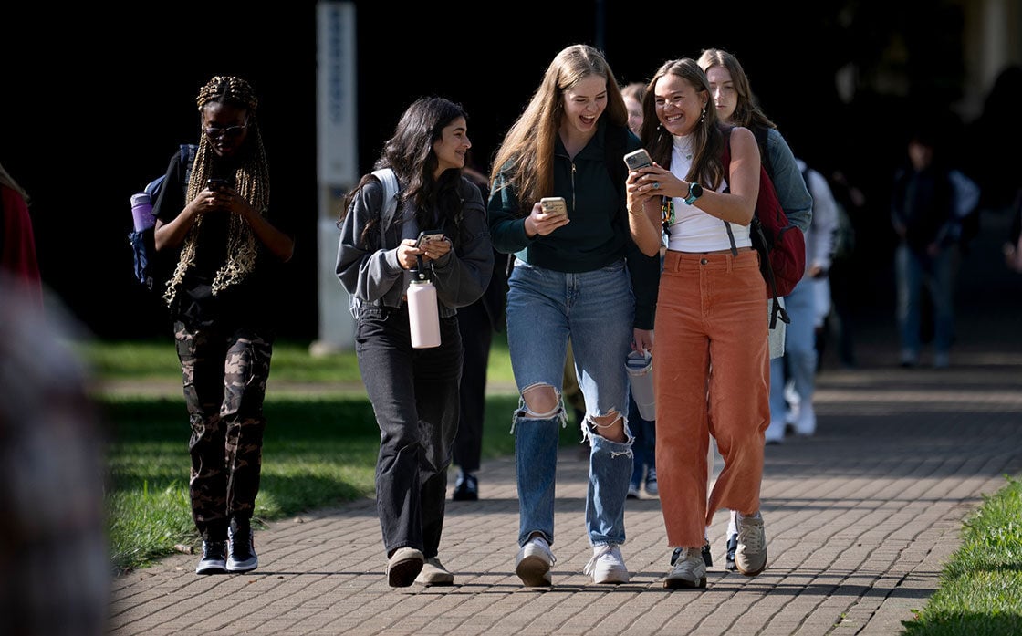 Four college students walking on campus, smiling and chatting, with two looking at a phone. The group is diverse, casually dressed, and enjoying a sunny day. Four college students walking on campus, smiling and chatting, with two looking at a phone. The group is diverse, casually dressed, and enjoying a sunny day.