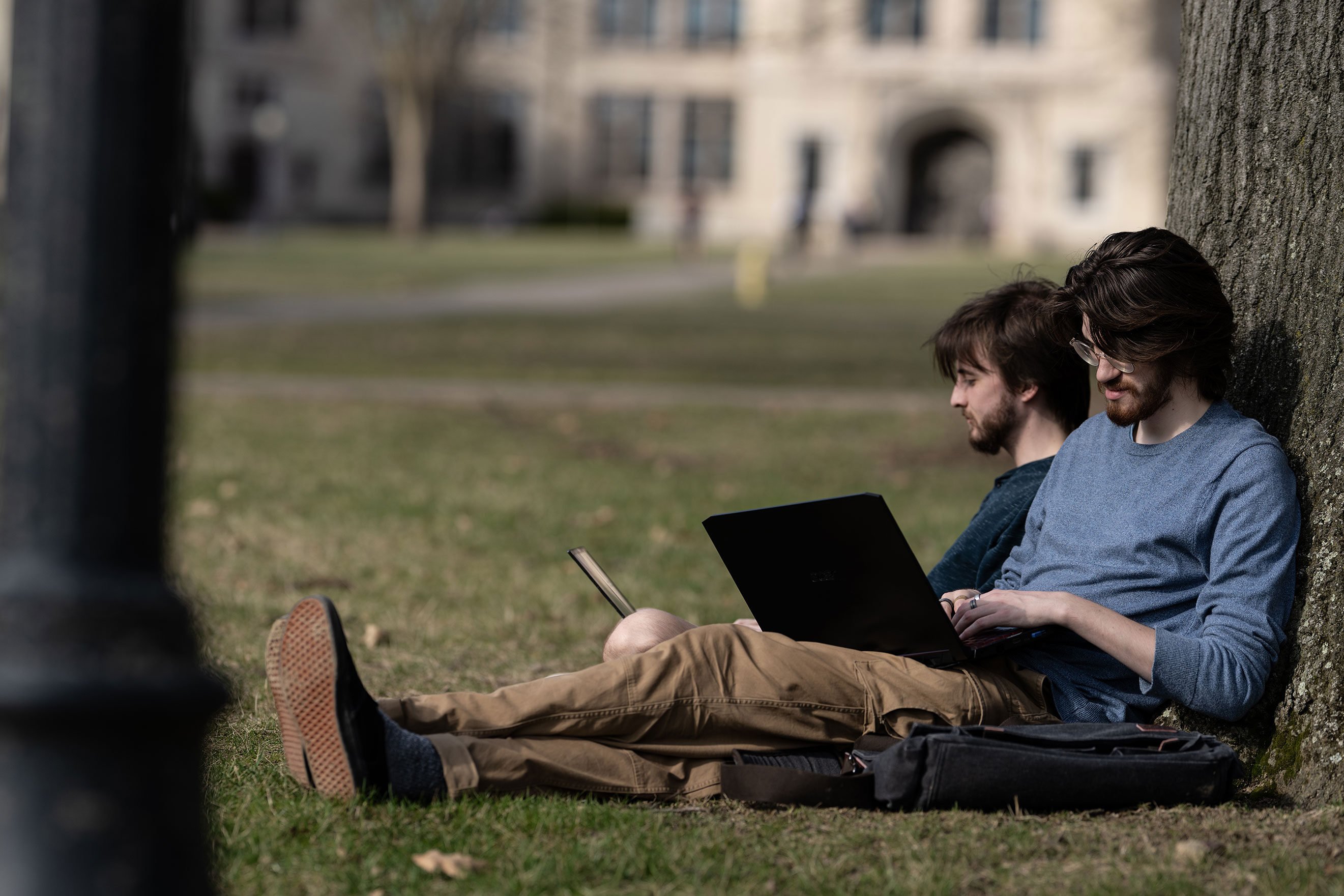 Two students sitting on grass under a tree on a college campus, each focused on their own device. One is reading from a tablet, while the other types on a laptop. Both are wearing casual clothes, with backpacks beside them, and a building with arched windows is visible in the background.